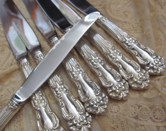 May include: A set of eight silver-plated dinner knives with ornate handles. The knives have a polished finish and are arranged in a fan shape.