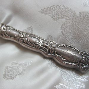 Op de afbeelding: Een zilverkleurige, sierlijke handgreep met bloemen- en bladerontwerpen. De handgreep heeft een gebogen vorm en een gedetailleerd patroon, met het woord "Patricia" gegraveerd. Het ligt op een witte, bedrukte stof.