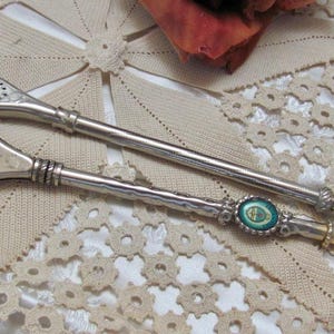 Könnte beinhalten: Zwei silberne Bombillas, traditionelle Yerba Mate Trinkhalme, liegen auf einer cremefarbenen Spitzendecke. Eine Bombilla hat einen ovalen türkisfarbenen und goldenen Akzent. Die andere hat ein schlichtes Design.