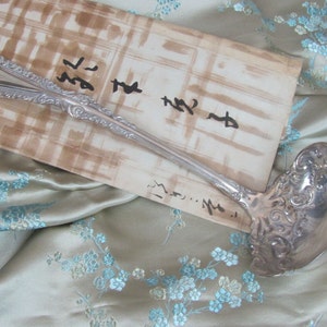 May include: A silver ladle with an ornate handle and a bowl with a decorative design. The ladle is lying on a piece of fabric with a floral pattern.