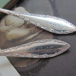 May include: Two antique silver spoons with ornate, engraved handles. The spoon bowls have the word "Souvenir" engraved in cursive. The spoons are set against a vintage background with a sepia-toned image.