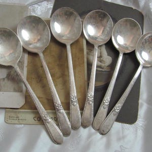 May include: Set of six silver-plated soup spoons with ornate handles. The spoons are arranged on a brown surface with a vintage photograph in the background.