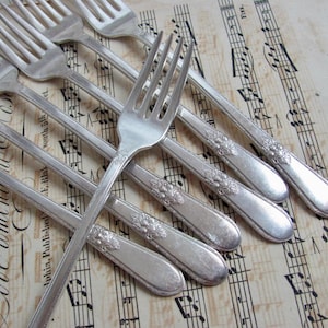 May include: A collection of silver forks with decorative floral patterns on the handles, arranged on a vintage sheet of music. The sheet music has faded text and musical notation, creating an antique and elegant feel.