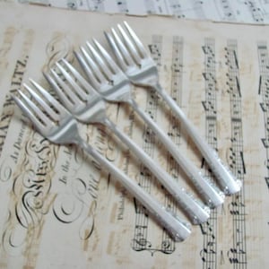 May include: Four silver forks with detailed handles are arranged diagonally on a vintage sheet music background. The sheet music has faded text and musical notes. The forks are clean and reflect light.