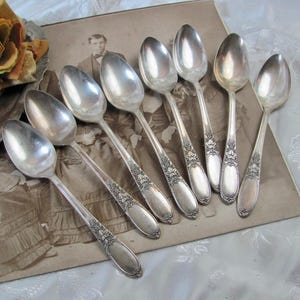 May include: A collection of silver spoons with ornate floral designs on the handles. The spoons are arranged on a vintage photograph, with a faded floral arrangement in the background. The spoons have a shiny, reflective surface.