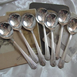 Puede incluir: Conjunto de ocho cucharas de sopa plateadas con mangos ornamentados. Las cucharas están dispuestas en forma de abanico sobre una superficie marrón.
