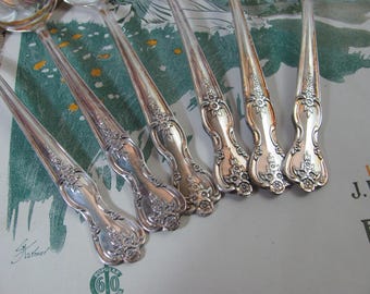 May include: A set of five silver-toned metal spoons featuring ornate floral embossed detailing on the handles. The spoons are arranged in a fan-like pattern, showcasing the intricate scrollwork and decorative motifs on the stems and ends.