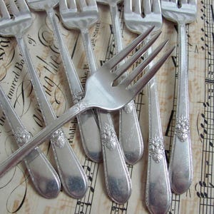 May include: Several antique silver forks with decorative floral patterns on the handles. The forks are displayed on a backdrop of aged sheet music, giving a classic, elegant appearance. The tines of the forks are clearly visible.