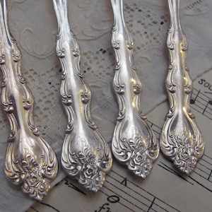 Può includere: Quattro cucchiai d'argento decorati con un motivo floreale. I cucchiai sono disposti su un tovagliolo di pizzo bianco con un foglio di musica sullo sfondo.