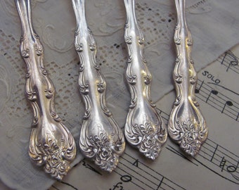 May include: Four ornate silver spoons with a floral design. The spoons are arranged on a white lace tablecloth with a sheet of music in the background.