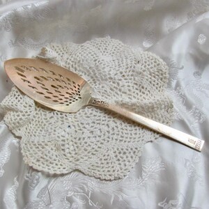May include: A silver-coloured slotted serving spoon with a long handle rests on a white, crocheted doily. The spoon has a decorative handle and a slotted, oval-shaped head. The background is a white, patterned fabric.
