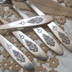 May include: A collection of silver forks with ornate black etched designs on the handles. The forks are arranged on a cream-colored surface with a decorative lace doily.