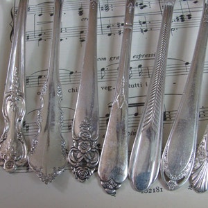 Puede incluir: Un juego de ocho cucharaditas plateadas con diseños ornamentados. Las cucharitas están dispuestas en fila sobre una hoja de música.