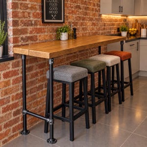 May include: A tall rectangular wooden bar table with a thick natural wood grain top supported by black industrial pipe legs. Four square bar stools with black metal frames and upholstered seats in grey, olive green, beige, and burnt orange fabric.