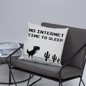 Puede incluir: Un cojín cuadrado blanco con el texto "NO INTERNET TIME TO SLEEP" en fuente pixelada negra. También se muestra un dinosaurio y cactus negros pixelados. El cojín está sobre una silla gris.