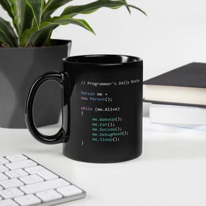 Puede incluir: Taza de café negra con texto blanco que dice "// Programmer's Daily Routine" y código en diferentes colores. El código dice "Person me = new Person(); while (me.Alive) { me.WakeUp(); me.Eat(); me.DoCode(); me.DebugMood(); me.Sleep(); }"