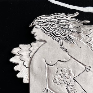 May include: A silver sculpture of an angel with wings outstretched, holding a bouquet of flowers. The angel is set against a black background with white paint splatters.