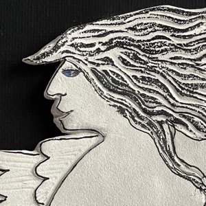 May include: A white ceramic sculpture of a woman with long black hair and a blue eye. The woman is depicted with wings and a flower on her chest.