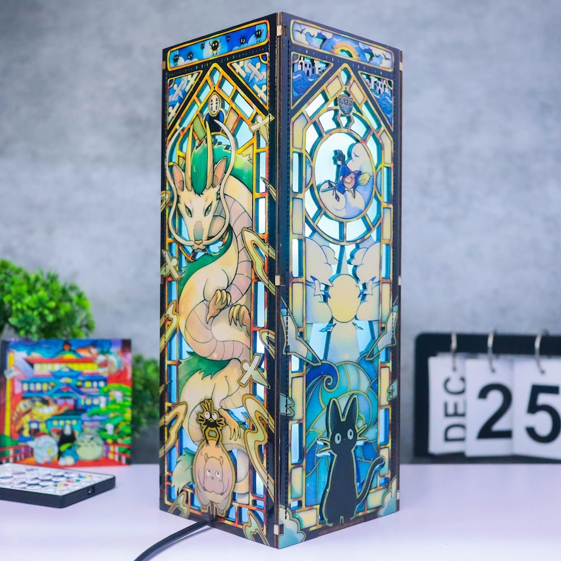 May include: A tall, rectangular wooden lamp featuring intricate, multi-layered laser-cut panels with stained glass-style illustrations of a long, serpentine dragon, a black cat, and various fantasy characters in shades of blue, green, and yellow.
