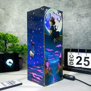 May include: A wooden night light featuring a whimsical design with a girl on a broomstick in front of a full moon. The lamp has a blue and purple color scheme with a starry night sky and city scene.