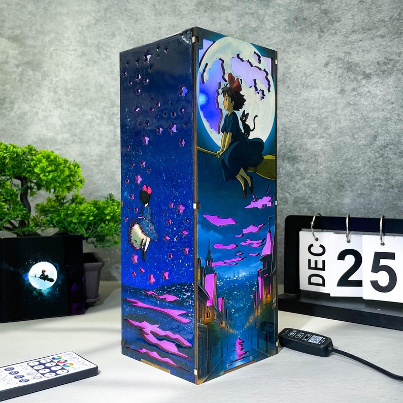 May include: A wooden night light featuring a whimsical design with a girl on a broomstick in front of a full moon. The lamp has a blue and purple color scheme with a starry night sky and city scene.