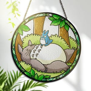 Może przedstawiać: Okrągły witraż przedstawiający scenę z filmu animowanego Mój sąsiad Totoro. Grafika przedstawia Totoro i mniejszego niebieskiego Totoro w leśnej scenerii, z zielonymi liśćmi i brązowymi pniami drzew. Panel ma czarną obwódkę i łańcuszek do zawieszenia.