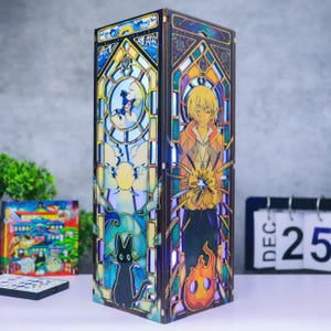 May include: A tall, rectangular wooden lamp featuring stained glass-style panels. One side depicts a blonde character with a fire spirit, while the other shows a black cat, a blue sky, and clouds. The lamp emits a soft, internal glow through the panels.