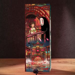 Puede incluir: L&aacute;mpara rectangular con una colorida ilustraci&oacute;n de una pel&iacute;cula de animaci&oacute;n. La obra de arte representa un puente, edificios y personajes con un estilo vibrante. La l&aacute;mpara tiene una base negra y un cable.