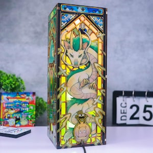May include: A rectangular wooden lamp featuring a stained glass-style design with a central green and white dragon illustration. The lamp emits a warm yellow glow and includes smaller character figures at the base and top, framed by dark wood panels.
