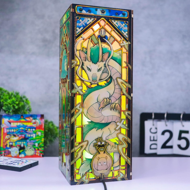 May include: A rectangular wooden lamp featuring a stained glass-style design with a central green and white dragon illustration. The lamp emits a warm yellow glow and includes smaller character figures at the base and top, framed by dark wood panels.