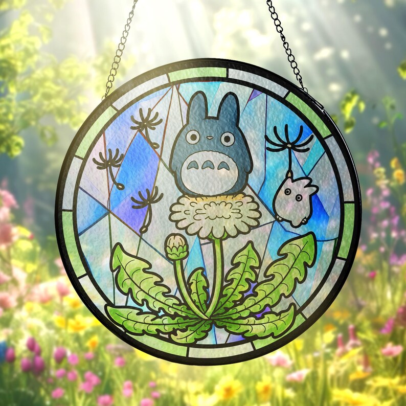 May include: Round stained glass window featuring a blue Totoro character and a small white creature on a dandelion. The window has a black border and is suspended by a chain. The background is a blurred image of a garden.