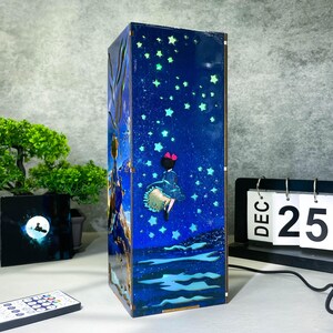 May include: A rectangular wooden lamp with a blue starry night design. The lamp features a girl with a red bow and a flowing dress. The lamp has a remote control and a calendar showing December 25th.