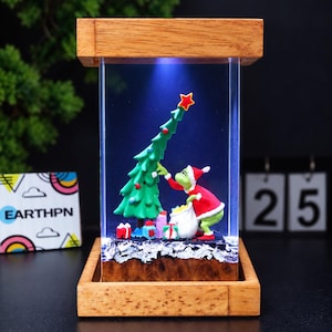 Könnte beinhalten: Ein rechteckiges Holzdisplay mit einem klaren Acrylzentrum, das eine grüne Figur in einem roten Anzug zeigt, die einen grünen Weihnachtsbaum mit einem roten Stern schmückt. Die Szene enthält kleine Geschenkkartons und einen weißen Sack, beleuchtet durch blaues Licht.