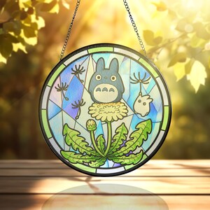 May include: Round stained glass window featuring a cartoon Totoro character on a dandelion. The artwork includes a blue, green, and yellow color palette, with a black border and chain for hanging. The background is a mix of blue and purple.