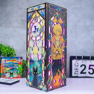 May include: A tall, rectangular wooden lamp featuring intricate, multi-colored stained glass-style panels. One side displays a blonde character holding a glowing light, while the other side depicts a black cat and a small figure in a blue hat.