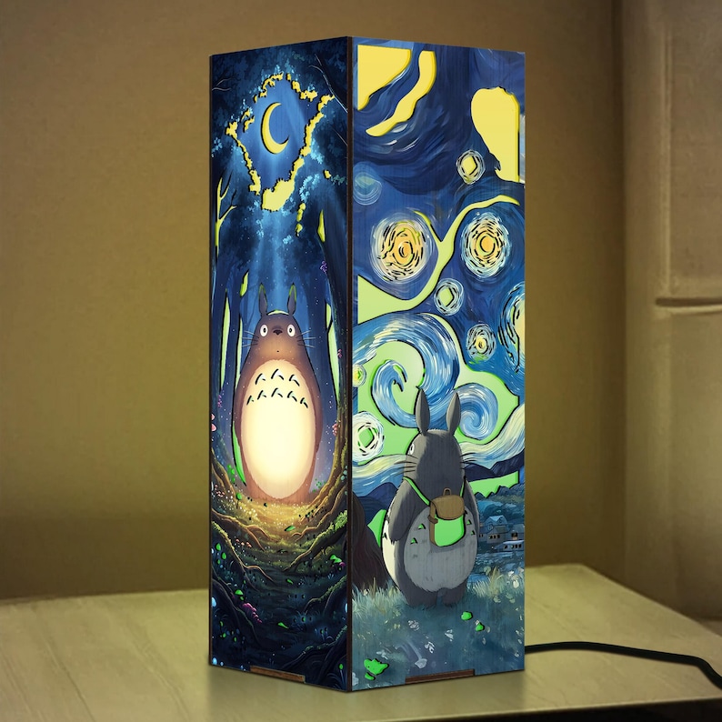 Może przedstawiać: Prostokątna drewniana lampa z ilustracjami inspirowanymi "Moim sąsiadem Totoro" studia Ghibli i "Gwiaździstą nocą" Van Gogha. Jedna strona przedstawia Totoro w lesie, druga scenę gwiaździstej nocy z postacią Totoro. Lampa stoi na drewnianej powierzchni.
