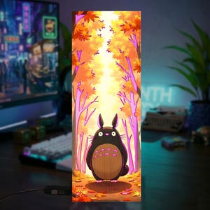 Puede incluir: L&aacute;mpara rectangular de madera con una escena de bosque de dibujos animados. La obra de arte representa una criatura negra con vientre marr&oacute;n en un bosque con hojas naranjas y amarillas. La l&aacute;mpara est&aacute; encendida, creando un brillo c&aacute;lido.