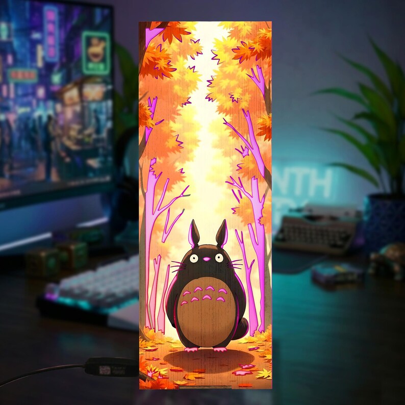 Puede incluir: L&aacute;mpara rectangular de madera con una escena de bosque de dibujos animados. La obra de arte representa una criatura negra con vientre marr&oacute;n en un bosque con hojas naranjas y amarillas. La l&aacute;mpara est&aacute; encendida, creando un brillo c&aacute;lido.