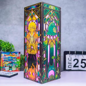 May include: A rectangular wooden lamp featuring intricate laser-cut panels with colorful stained-glass style illustrations. One side depicts a blonde character with a flame, while the other shows a green creature and foliage in a gothic arch design.