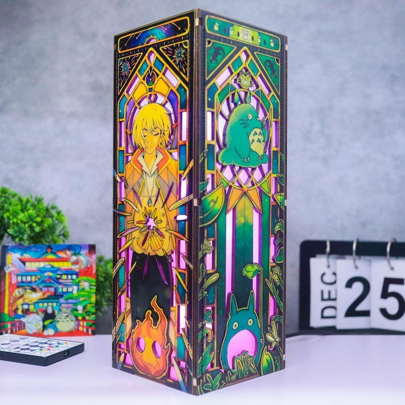 May include: A rectangular wooden lamp featuring intricate laser-cut panels with colorful stained-glass style illustrations. One side depicts a blonde character with a flame, while the other shows a green creature and foliage in a gothic arch design.