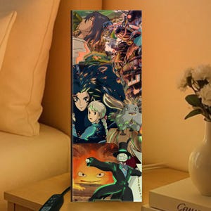 K&ouml;nnte beinhalten: Rechteckige Holzlampe mit einer Collage von Anime-Charakteren und -Szenen. Das Kunstwerk zeigt Charaktere mit langen Haaren, ein junges M&auml;dchen und eine Figur in einem schwarzen Anzug. Die Lampe ist beleuchtet und strahlt ein warmes Licht aus.