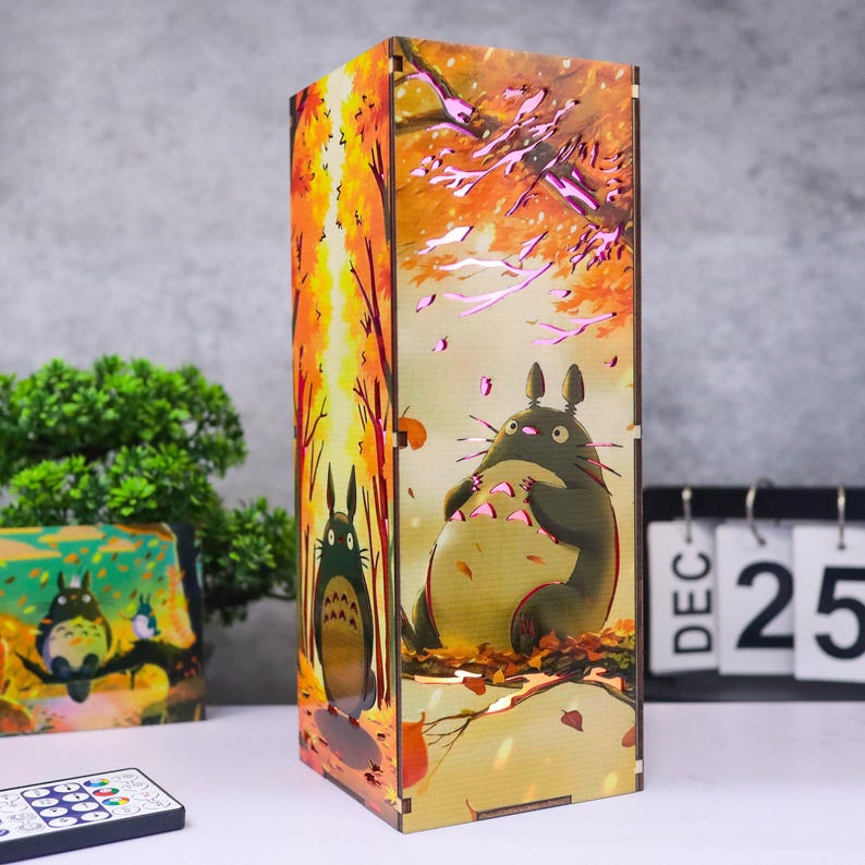 Puede incluir: L&aacute;mpara rectangular de madera con una escena de bosque y dos personajes de Totoro. La l&aacute;mpara emite una luz c&aacute;lida y la obra de arte muestra una escena oto&ntilde;al con hojas naranjas y amarillas. Se ve un mando a distancia. La l&aacute;mpara mide unos 20 cm de altura.