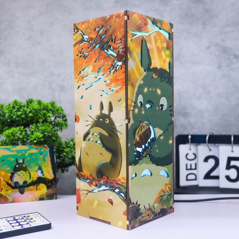 Puede incluir: L&aacute;mpara rectangular de madera con una ilustraci&oacute;n de dibujos animados de los personajes de Totoro en una escena de bosque con colores oto&ntilde;ales. La l&aacute;mpara emite un brillo c&aacute;lido.