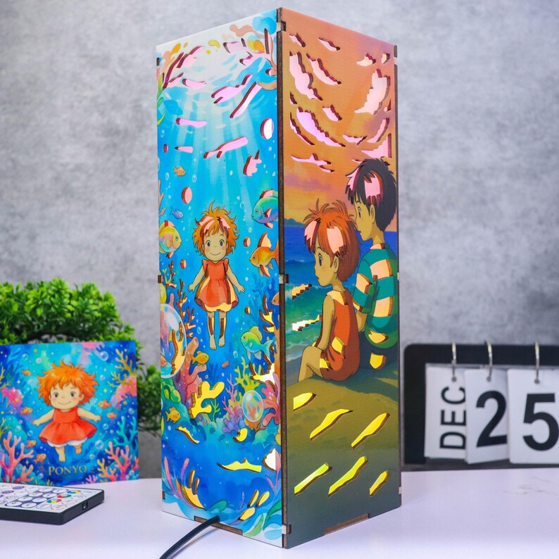 May include: A rectangular wooden lamp featuring colorful illustrations of characters from Ponyo. One side shows a character underwater with coral and fish, while the other side depicts two characters sitting by the sea at sunset with orange clouds.