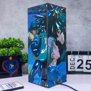 Op de afbeelding: Een rechthoekige houten lamp met een blauw en blauwgroen anime-stijl illustratie van twee paren die elkaar omarmen. De lamp heeft een zwart netsnoer en staat naast een afstandsbediening en een kalender met 25 december.