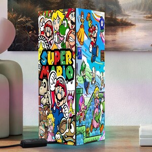 Puede incluir: Una l&aacute;mpara rectangular con un colorido collage de personajes de Super Mario y el logotipo del juego. El dise&ntilde;o de la l&aacute;mpara incluye a Mario, Luigi, la Princesa Peach y otros personajes ic&oacute;nicos. La l&aacute;mpara est&aacute; sobre una superficie de madera.