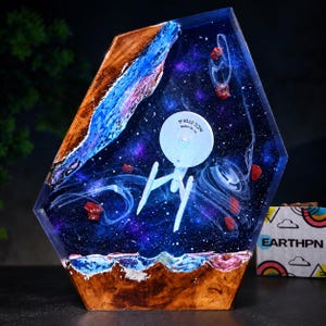 May include: A resin art piece featuring a miniature NCC-1701-A starship model suspended in a deep blue and purple space scene with swirling white nebula clouds and red asteroid fragments, framed by natural wood edges at the top and bottom.