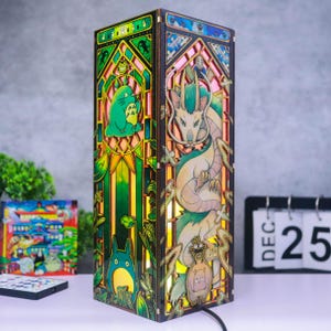 May include: A rectangular wooden lamp featuring intricate laser-cut panels with colorful stained-glass style illustrations. One side displays a green forest creature, while the other side depicts a long, serpentine dragon figure with light emanating from within.