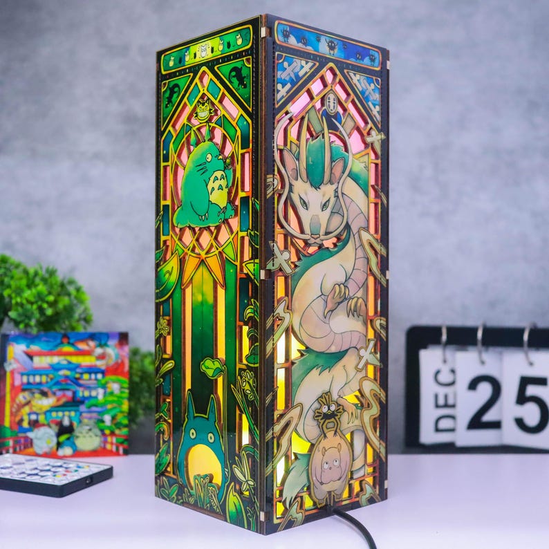 May include: A rectangular wooden lamp featuring intricate laser-cut panels with colorful stained-glass style illustrations. One side displays a green forest creature, while the other side depicts a long, serpentine dragon figure with light emanating from within.