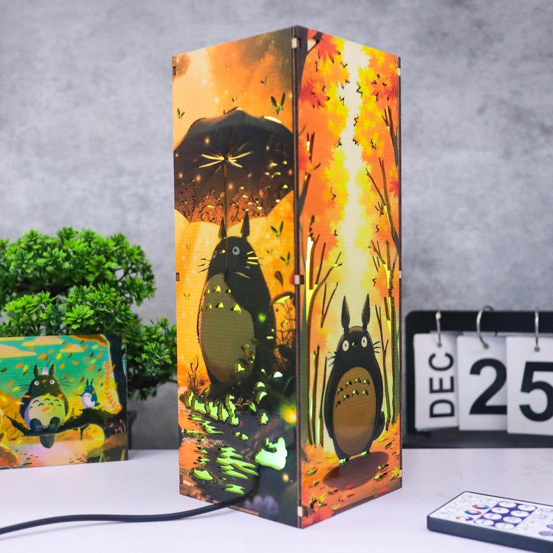 Puede incluir: L&aacute;mpara decorativa de madera con dise&ntilde;o de personaje de dibujos animados. La l&aacute;mpara presenta una escena de bosque oto&ntilde;al naranja y amarillo con un personaje sosteniendo un paraguas. La l&aacute;mpara es rectangular y tiene un cable de alimentaci&oacute;n negro.
